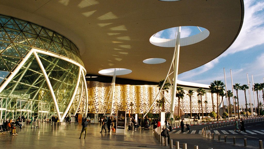 Façade moderne de l’aéroport de Marrakech avec sa structure géométrique, ses ouvertures circulaires et des voyageurs devant l’entrée.