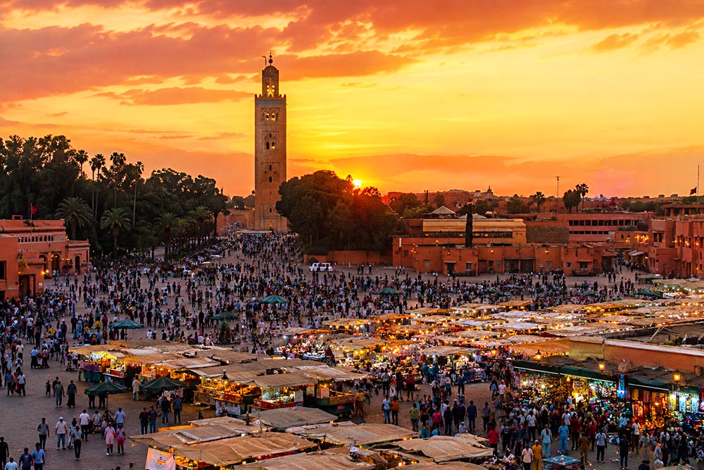 Place Jemaa el-Fna au coucher du soleil, animée par la foule et les food stalls, avec la Koutoubia en arrière-plan.